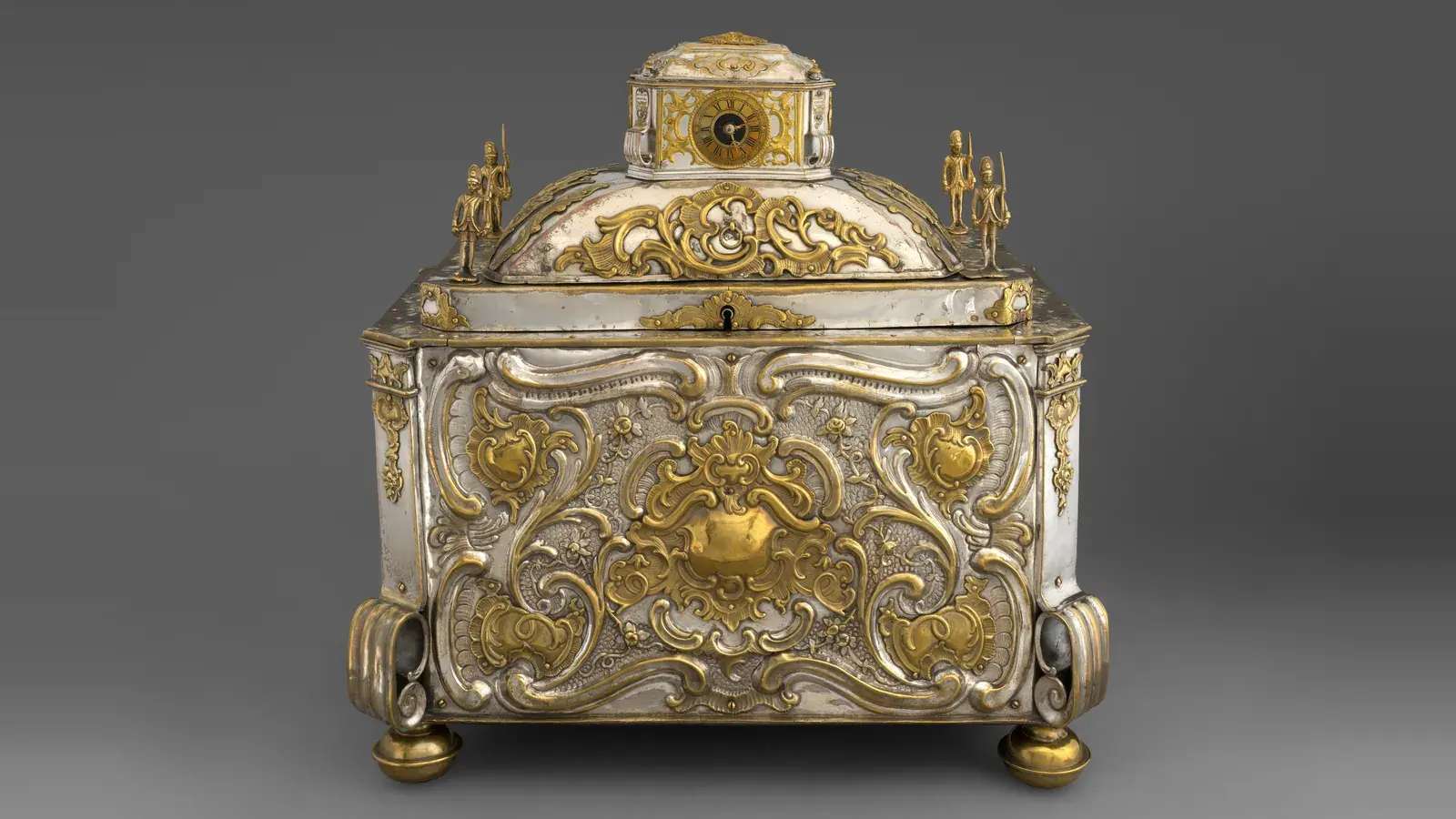 An ornate 1750 strongbox in silver and gold tones, a small clock set into its domed lid above the keyhole