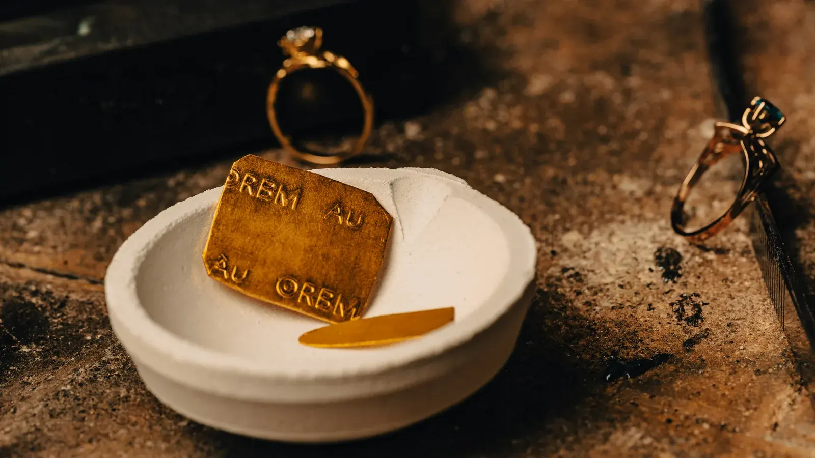 A poured gold ingot stamped Au resting in a ceramic crucible dish, two gem-set rings on the bench beside it.
