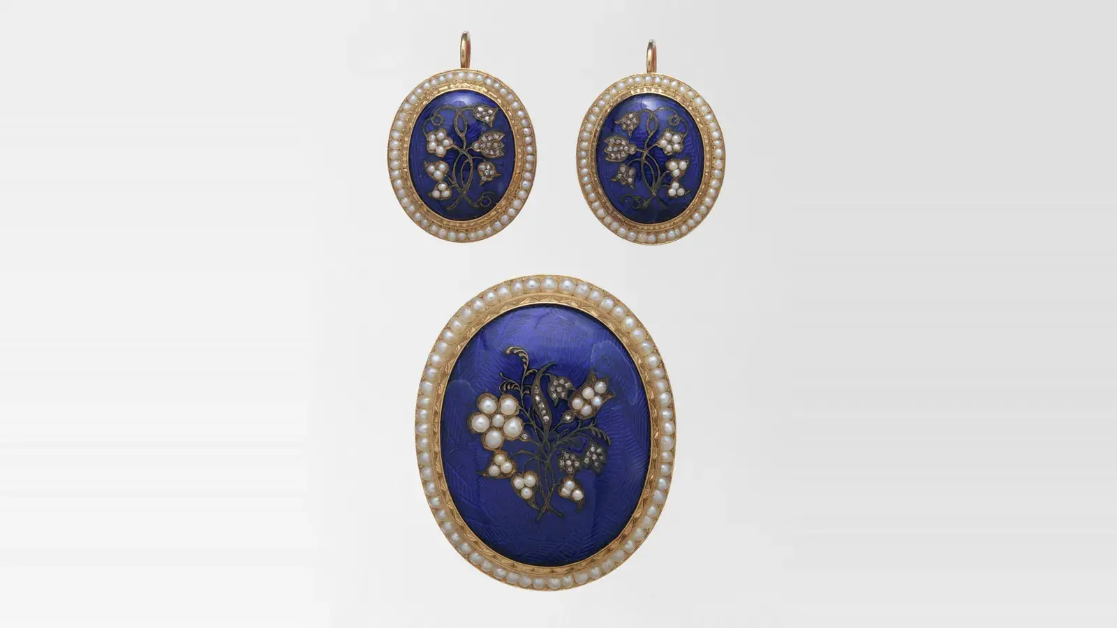 A matching brooch and pair of earrings in blue enamel, each framed by seed pearls around a spray of pearls and small diamonds, laid out together on grey